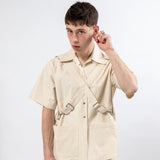 Model wearing the Emily Millichip Bondage Shirt in natural seeded cotton canvas, with crossed buckle straps and large patch pockets against a white studio background.