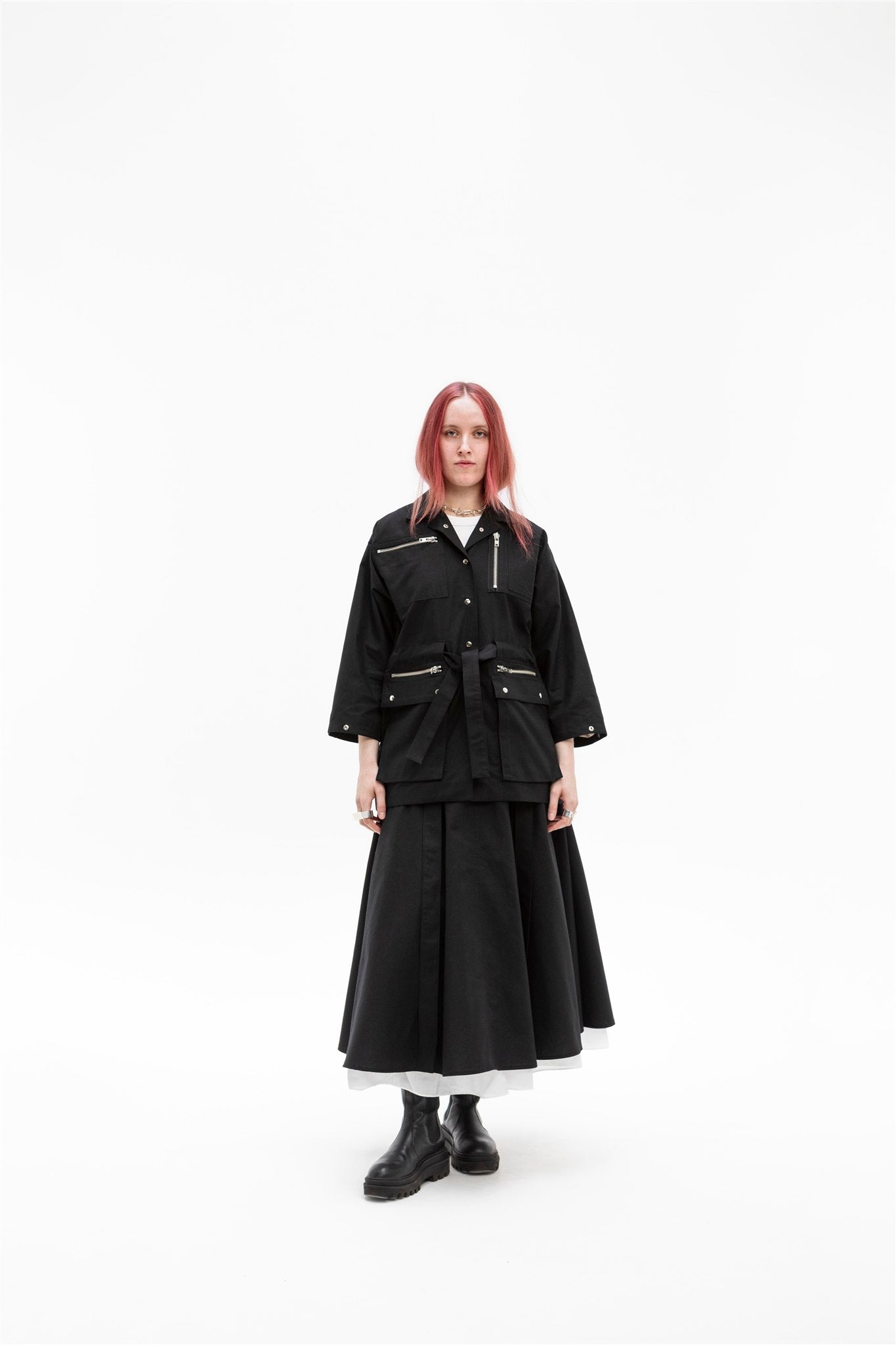 Full-length front view of the Emily Millichip Gravity Field Jacket in black heavyweight sanded canvas, worn with  relaxed boxy fit, zipped utility pockets, styled with a matching long black skirt.