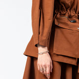 Side detail of the Emily Millichip Gravity Field Jacket in bronze heavyweight sanded canvas, showing the cinched waist, cropped sculptural sleeve, black oxidised metal zips, and oversized utility pockets with black press studs.