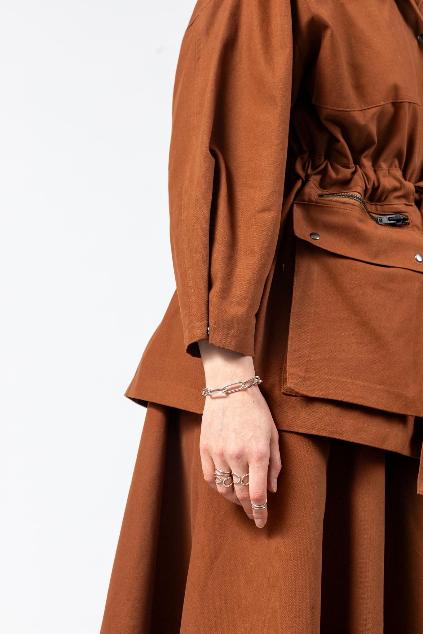 Side detail of the Emily Millichip Gravity Field Jacket in bronze heavyweight sanded canvas, showing the cinched waist, cropped sculptural sleeve, black oxidised metal zips, and oversized utility pockets with black press studs.