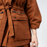 Close-up of the Emily Millichip Gravity Field Jacket in bronze heavyweight sanded canvas, showing the cinched waist with fabric tie belt, zipped pockets with black oxidised metal zips, oversized utility pockets with black press studs, and sculptural sleeves.