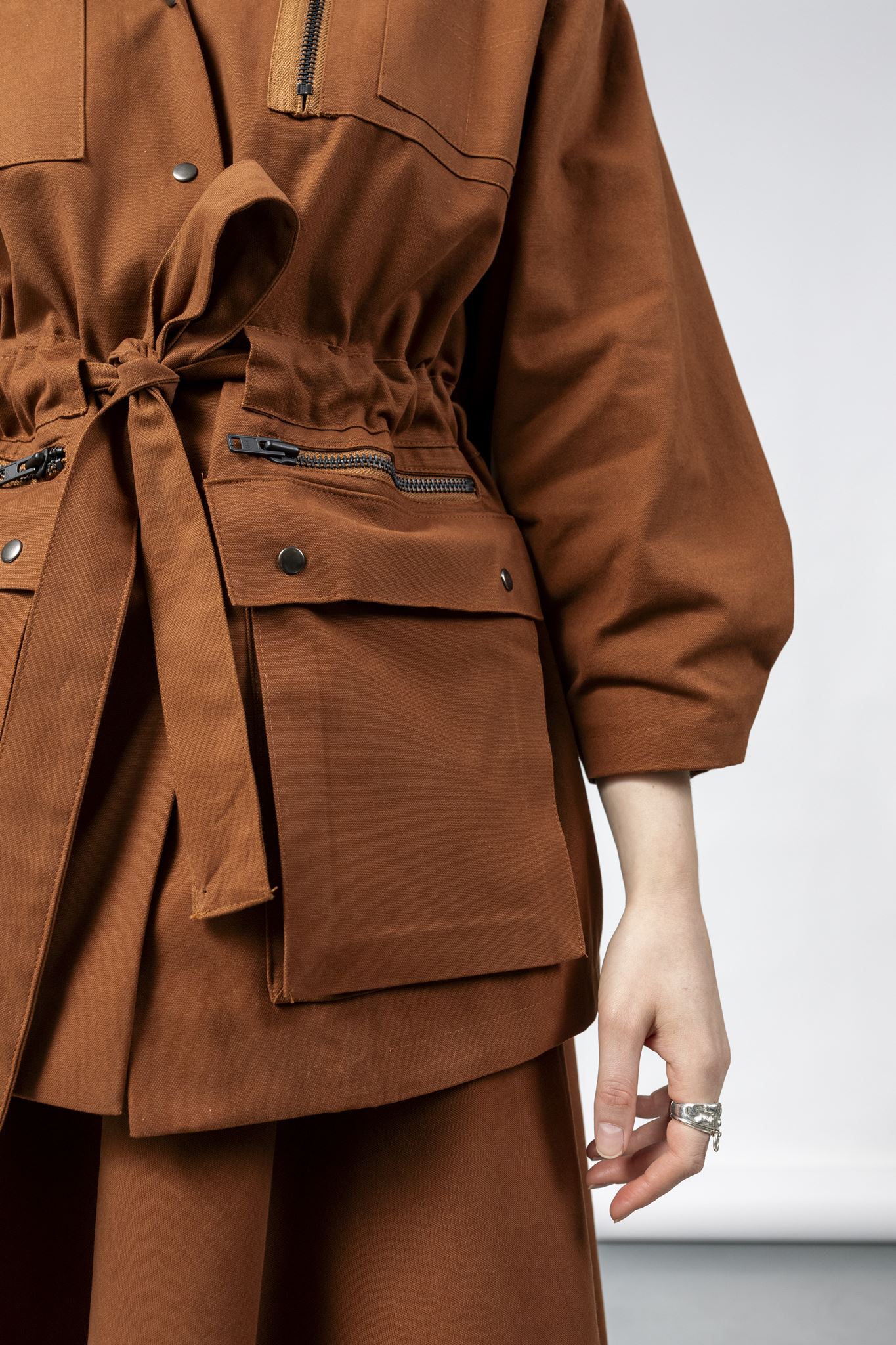 Close-up of the Emily Millichip Gravity Field Jacket in bronze heavyweight sanded canvas, showing the cinched waist with fabric tie belt, zipped pockets with black oxidised metal zips, oversized utility pockets with black press studs, and sculptural sleeves.